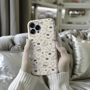 May include: A beige phone case featuring a repeating pattern of hockey-related illustrations. The design includes hockey sticks, skates, helmets, pucks, and hearts. The case is held by a person wearing a white sweater.