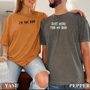 May include: A mustard yellow t-shirt with the black text I'M THE BOO and a charcoal gray t-shirt with the white text JUST HERE FOR MY BOO. Both shirts feature short sleeves, a crew neckline, and a relaxed fit with a distressed, vintage-style finish.
