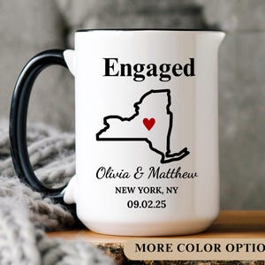 May include: White ceramic mug with a black handle and rim. The mug features the word "Engaged" above a black outline of New York State with a red heart. Below the state outline are names and a date.