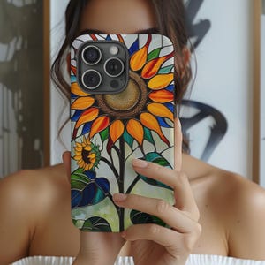 May include: A smartphone case featuring a stained-glass style sunflower design. The case showcases a large, detailed sunflower with orange, yellow, and blue petals, a brown center, and green leaves. The design is set against a light background.