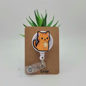 May include: A white retractable badge holder with a cartoon orange cat design. The holder is attached to a brown card with the text "Hold My Badge".