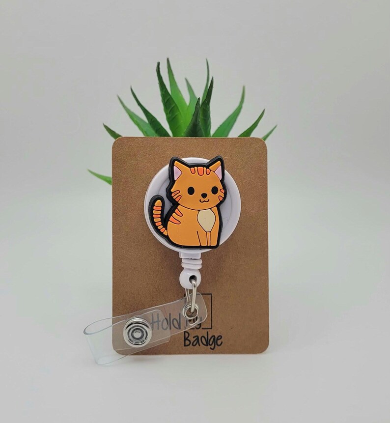 May include: A white retractable badge holder with a cartoon orange cat design. The holder is attached to a brown card with the text "Hold My Badge".