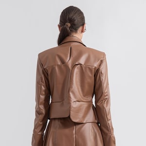 May include: A brown leather jacket and matching skirt set. The jacket has a tailored fit with a notched collar and a cropped hem. The skirt is a mini skirt. The outfit is paired with brown knee-high platform boots.