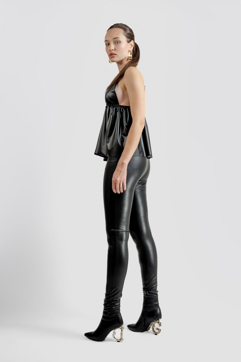 May include: A woman in a black halter top, leggings, and ankle boots. The boots have a gold decorative heel. The outfit is made of a shiny, leather-like material. The background is plain white.