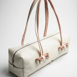 Puede incluir: Bolso rectangular color crema con cierre de cremallera y dos juegos de asas. Las asas son rosas y blancas, y el bolso tiene lazos rosas en cada extremo. El bolso est&aacute; hecho de un material texturizado.