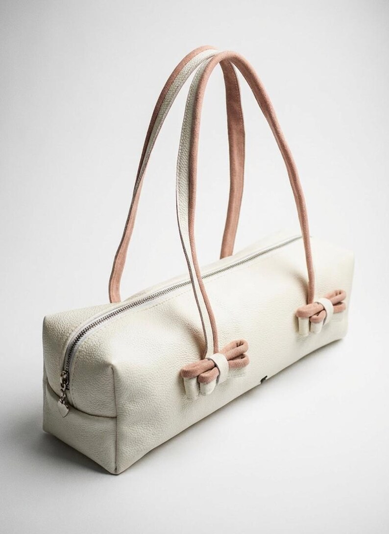 Puede incluir: Bolso rectangular color crema con cierre de cremallera y dos juegos de asas. Las asas son rosas y blancas, y el bolso tiene lazos rosas en cada extremo. El bolso est&aacute; hecho de un material texturizado.