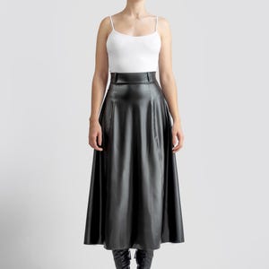 May include: A black leather midi skirt with a high waist and a flared silhouette. The skirt is worn with a white tank top and black boots.