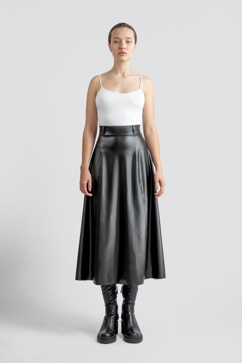 May include: A black leather midi skirt with a high waist and a flared silhouette. The skirt is worn with a white tank top and black boots.