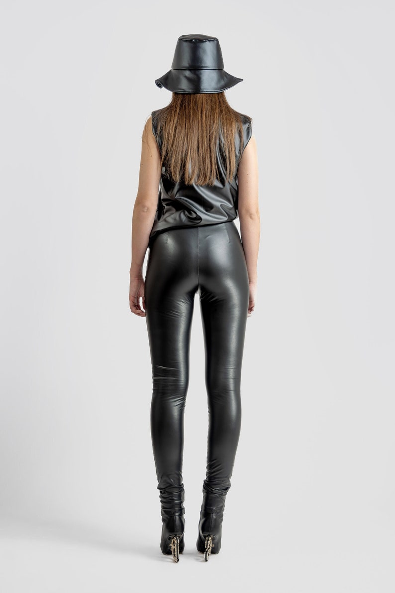 May include: A person wearing a black leather bucket hat, sleeveless top, and leggings. The outfit is all black and appears to be made of a shiny, faux leather material. The person is also wearing black boots.