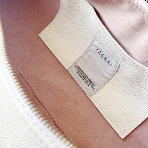 Puede incluir: Primer plano del interior de un bolso de cuero color crema con forro rosa claro. Una etiqueta de cuero blanco con la marca "TACRAI" e informaci&oacute;n de derechos de autor es visible. El bolso tiene una cremallera.