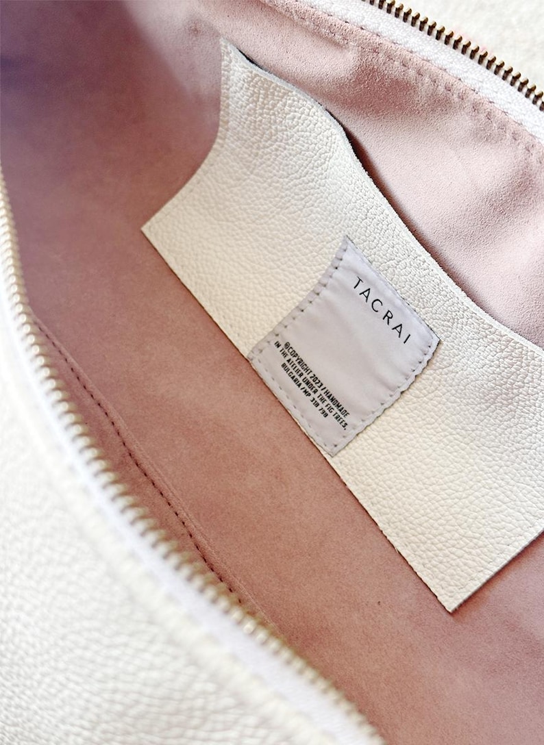 Puede incluir: Primer plano del interior de un bolso de cuero color crema con forro rosa claro. Una etiqueta de cuero blanco con la marca "TACRAI" e informaci&oacute;n de derechos de autor es visible. El bolso tiene una cremallera.