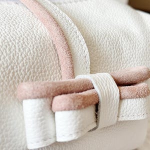 Puede incluir: Primer plano de un bolso de cuero blanco con una superficie texturizada. El bolso presenta un acento de ante rosa en el asa y un lazo decorativo. El lazo est&aacute; hecho de cuero blanco con ribete de ante rosa.