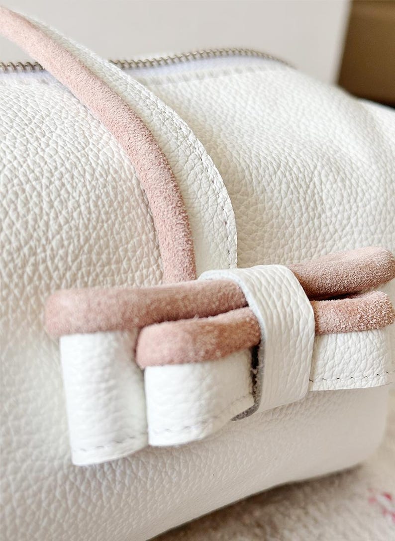 Puede incluir: Primer plano de un bolso de cuero blanco con una superficie texturizada. El bolso presenta un acento de ante rosa en el asa y un lazo decorativo. El lazo est&aacute; hecho de cuero blanco con ribete de ante rosa.