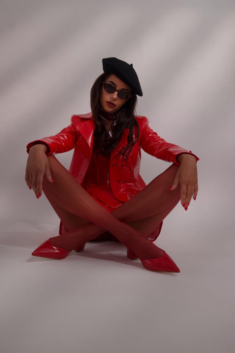 May include: A woman wearing a red leather jacket, red tights, and red heels sits with her legs crossed. She is wearing a black beret and sunglasses.