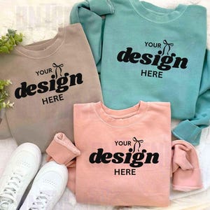 Puede incluir: Tres sudaderas de cuello redondo en beige, verde azulado y coral, cada una con el texto "YOUR design HERE" en negro. Zapatillas blancas en la parte inferior izquierda. Las sudaderas están sobre una superficie blanca texturizada.