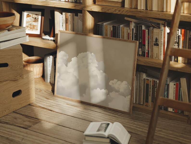 May include: A framed print of a cloudy sky with a light brown wooden frame. The print is leaning against a wooden bookshelf filled with books.