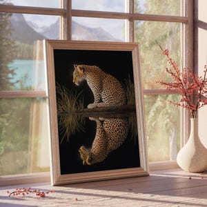May include: A framed print of a leopard sitting by a body of water, with its reflection visible in the water. The print is set against a dark background and features a gold-colored frame.