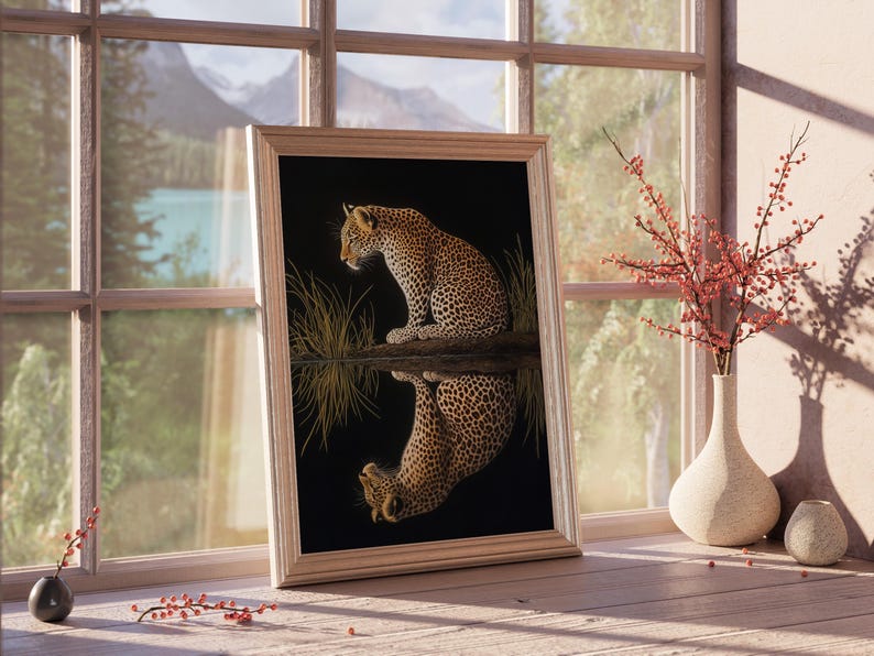 May include: A framed print of a leopard sitting by a body of water, with its reflection visible in the water. The print is set against a dark background and features a gold-colored frame.