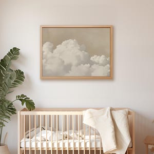 May include: A framed print of a beige and white cloud formation against a light brown background. The print is hanging on a white wall above a wooden crib with a white blanket.