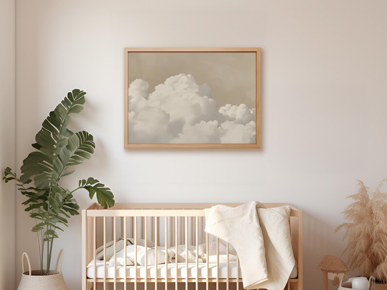 May include: A framed print of a beige and white cloud formation against a light brown background. The print is hanging on a white wall above a wooden crib with a white blanket.
