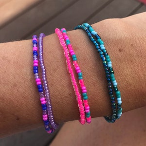 May include: Four beaded bracelets in different colors, including pink, blue, purple, and teal. The bracelets are stacked on a person's wrist.