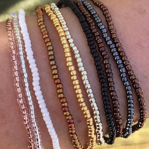 May include: A collection of beaded bracelets in various colors, including rose gold, silver, white, bronze, gold, black, and brown. The bracelets are made of small, round beads strung together, creating a delicate and stylish accessory. The bracelets are shown on a wrist.