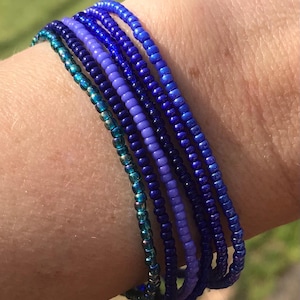May include: A collection of beaded bracelets in shades of blue and purple. The bracelets are made of small, round beads and are stacked together on a wrist. The colors range from teal to deep indigo, creating a gradient effect.