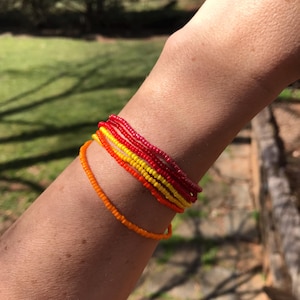 May include: A selection of colourful beaded bracelets in red, yellow, and orange hues. The bracelets are stacked on a wrist, set against a natural outdoor backdrop. The bracelets are made of small, round beads.