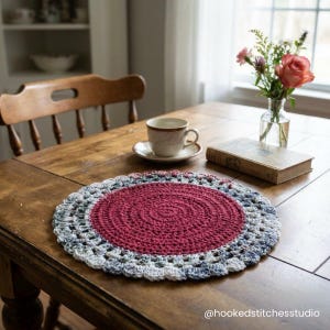 Op de afbeelding: Een handgemaakte, ronde gehaakte placemat met een dieprood midden en een veelkleurige rand van blauw, grijs en wit. De placemat ligt op een houten tafel, samen met een kop en schotel, een boek en een vaas met bloemen.