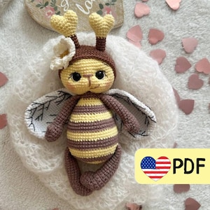 Könnte beinhalten: Häkel-Amigurumi-Bienenspielzeug mit braunen und gelben Streifen, weißen Flügeln und einer weißen Blume auf dem Kopf. Die Biene sitzt auf einer weißen, flauschigen Oberfläche mit rosa Herzen, die um sie herum verstreut sind. Der Text "PDF" ist in der unteren rechten Ecke des Bildes sichtbar.