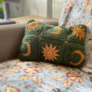 May include: A green crocheted pillow with a pattern of suns and moons in a light brown color. The pillow is sitting on a light blue throw blanket with a pattern of leaves and pumpkins.
