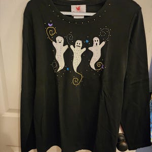 以下が含まれることがあります： 3つの笑顔のゴーストデザインが特徴の黒い長袖シャツ。ゴーストは白で、装飾的な渦巻きとドットが付いています。シャツは、小さな金色の装飾が施されたクルーネックです。