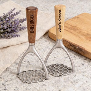 May include: Two potato mashers with wooden handles engraved with "John's Kitchen". The mashers have a silver-coloured metal base with a honeycomb pattern. One handle is light wood, the other is dark wood. A wooden chopping board and lavender sprigs are in the background.