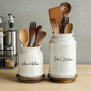 May include: Two white ceramic kitchen utensil holders with the text "John's Kitchen" in black script. The holders sit on wooden bases and contain various wooden spoons, forks, and spatulas. Kitchen accessories.