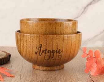 May include: Two wooden bowls stacked on top of each other. The bottom bowl has the name "Anggie" engraved on the front. The bowls are light brown with visible wood grain. The bowls are sitting on a wooden surface.