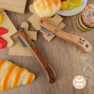 Op de afbeelding: Twee broodmessen met houten handvatten en gepersonaliseerde gravures. Het mes aan de linkerkant heeft een "G" gegraveerd op het handvat en het mes aan de rechterkant heeft een "M" gegraveerd op het handvat. De messen liggen op een houten oppervlak met gebak en fruit op de achtergrond.