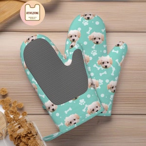 May include: A pair of teal blue oven mitts with a white background and a repeating pattern of a white dog's face and white bone shapes. The mitts have a gray silicone heat-resistant lining.