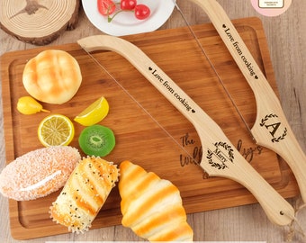 May include: Two wooden bread knives with wire blades on a wooden cutting board. The knives have engraved text that says "Love from cooking" and "The Willys". One knife has a wreath with the letter "A" inside and the other has a wreath with the name "Marry" inside. The cutting board has a variety of breads, fruit, and other food items on it.