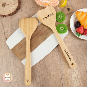 Könnte beinhalten: Zwei herzförmige Küchenlöffel aus Holz mit personalisierten Gravuren. Der erste Löffel trägt die Aufschrift "Jenny ♥ Jack" und der zweite Löffel trägt die Aufschrift "Sally's kitchen".