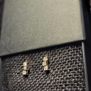 May include: A pair of gold-toned stud earrings, each featuring three square-cut, clear gemstones. The earrings are displayed inside a black jewelry box with a textured, dark gray interior.