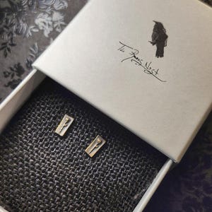 May include: Open white box with two small gold-colored rectangular earrings. The earrings are inside a box with the text "The Fae's Nest" and a bird illustration. The box is on a dark patterned surface.