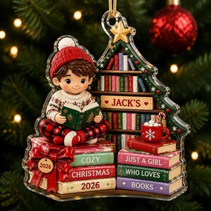 May include: A Christmas tree-shaped ornament featuring a child in a red beanie and patterned sweater reading a book. The design includes shelves of books, a red mug with marshmallows, and stacked books labeled "COZY CHRISTMAS 2026" and "JUST A GIRL WHO LOVES BOOKS".