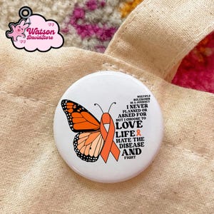 Puede incluir: Un botón blanco con un diseño de mariposa monarca y una cinta naranja. El texto en el botón dice: "La esclerosis múltiple es un viaje, nunca lo planeé ni lo pedí, pero elijo amar la vida y odiar la enfermedad y luchar."