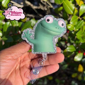 Puede incluir: Un portatarjetas de rana de dibujos animados verde con un borde de purpurina plateada. La rana tiene ojos rosas y está saludando. El portatarjetas tiene un cordón retráctil de plástico transparente y un clip metálico.