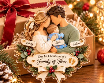 May include: A clear acrylic Christmas ornament featuring a family of three. The figures include a person with long blonde hair, a person with dark hair, and a baby in blue pajamas. Text reads: Kaisey, Jack, Wilson, and Our 1st Christmas as a Family of Three.