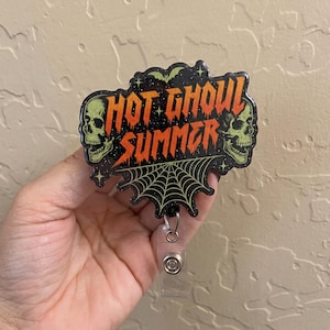 May include: A black badge reel featuring the text "HOT GHOUL SUMMER" in orange, stylized lettering. The design includes two pale green skulls on the sides, a green bat at the top, and a spiderweb graphic at the bottom, all set against a glittery background.