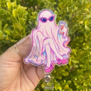 May include: An acrylic badge reel featuring a ghost illustration wearing pink cat-eye sunglasses and platform heels while holding a can of Diet Coke. The ghost is draped in a light pink sheet with blue outlines, sitting with legs crossed.