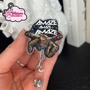 May include: A badge reel with a clear plastic body and a retractable cord. The badge features a graphic of a creature with the word "AMAZE" in white lettering. The background is black with glitter.