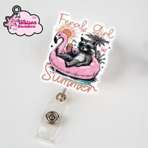 May include: A badge reel featuring a raccoon wearing sunglasses, relaxing on a pink flamingo floatie. The words "Feral Girl Summer" are written above the raccoon, with the word "Summer" below. The reel has a clear plastic strap and a metal clip.