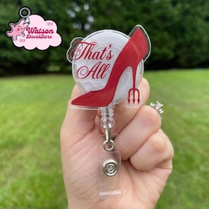 Puede incluir: Un carrete de insignia transparente con un diseño de zapato de tacón alto rojo y el texto "That's All". El carrete de la insignia también presenta un pequeño tridente y un clip de plástico transparente.